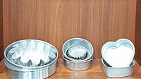 Image showing large fluted metal cake pan, small round pans stacked inside a larger pan, and two white heart-shaped cake pans side by side on a wooden shelf.