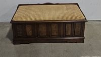Wooden padded cedar chest bench with decorative panels on front base and beige cushioned top with dark trim.