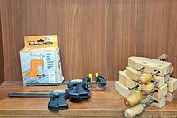 All clamps displayed together on a wooden surface with a wood backdrop, showing two mini bar clamps, the boxed hold down clamp, and four wooden clamps.