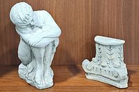 Photo showing sitting boy concrete sculpture next to hanging concrete pillar column piece against wooden background.