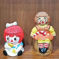Front view of both vintage Raggedy Ann ceramic cookie jars placed side by side on a wooden surface with plain wooden background.