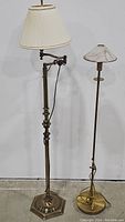 Two vintage brass floor lamps side by side against a plain white wall on concrete floor. Left lamp has a pleated cream shade and swing arm. Right lamp is slender brass with frosted glass shade.
