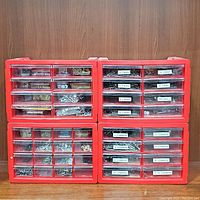 Four red hardware storage caddies filled with assorted screws, nails, and fasteners organized in clear plastic drawers, arranged on a wooden shelf