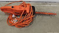 Top view of Black & Decker electric hedge trimmer laying on concrete, showing the orange trimmer body and long orange power cord coiled on top.