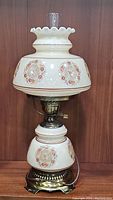 Full front view of vintage hurricane parlor table lamp against wood wall showing scalloped top glass shade with floral medallions, middle glass body with matching painted decorations, and brass base and fittings.