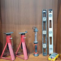 Photo showing two red adjustable jack stands, Black & Decker Bright Bar, two levels (one black, one white), and Fluke multimeter with test leads on wooden surface against wood panel background.