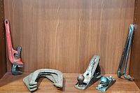 Pipe wrench, C-clamp, block planes, crowbars, and horseshoes displayed on wood shelf