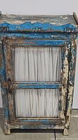 View of rustic cabinet front showing door with glass pane and curtain, distressed blue paint and scalloped decorative top edge.