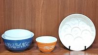 Vintage Pyrex mixing bowls: large blue Colonial Mist 403 bowl, orange 401 brown wheat pattern bowl, and white molded egg tray, all arranged on wooden surface.