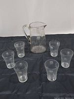 Set of one clear glass pitcher surrounded by six matching clear etched glass tumblers on dark surface