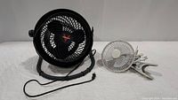 Photo of both fans side by side on a white surface: black Vornado floor fan and small white clamp fan with clip.