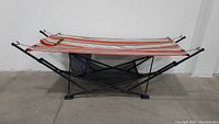 Foldable portable hammock with rust striped fabric, pillow and black storage bag on bottom, metal frame black.