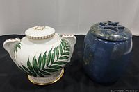 Front view of two large lidded ceramic jars: white with palm leaf and gold trim by Boehm, and blue glazed stoneware unbranded jar
