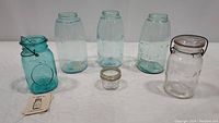 Six Mason jars on white surface including blue-green and clear glass jars, one with wire bail lid and one small jar with metal lid.