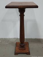 Full view of the antique neoclassical pedestal table showing square top, carved pedestal column, and square base on concrete floor.