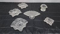 Several pressed glass pieces displayed on a black cloth: 3 diamond hobnail Westmoreland dishes with ruffled sawtooth rims, 1 glass top hat, 1 square trinket dish, 1 fan shaped serving dish.