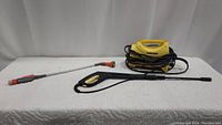 Yellow Karcher electric pressure washer with black hose coiled around unit and long spray gun placed in front, grey and orange garden hose nozzle extension lying nearby on white surface.