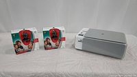 Two Logitech QuickCam Communicate STX webcams in unopened boxes displayed next to an HP Photosmart All-In-One printer/copier on a white background.