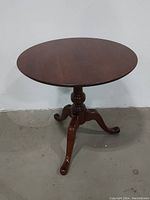 Full side view of the round mahogany tilt top table showing the turned pedestal and three curved legs on concrete floor.