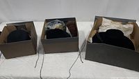 Three cardboard boxes each containing one or more black dress hats wrapped in tissue paper, showing overall lot packaging and condition.