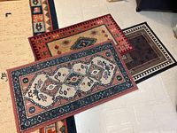 Three area rugs stacked and overlapping showing different tribal and geometric patterns and colors including red, cream, navy, and brown.