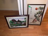 Photo shows both framed fabric art pieces leaning against the wall on wooden floor, one with peacocks and one with pagoda scene.