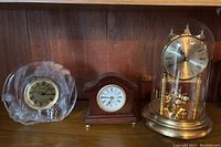 Three clocks on wood shelf: Birks dome clock with gold base and pendulum balls, small wood table clock with arched top, and Studios crystal clock with clear body and gold accents.