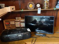 Sony CFD-10 CD radio cassette recorder and RCA 24-inch TV with remote on wooden shelf with cassette tapes behind.