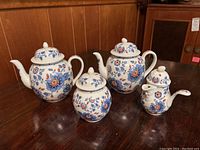 Two Estee Lauder porcelain teapots, creamer and sugar bowl with blue and orange floral pattern on white porcelain, displayed on wooden surface against a wood-paneled wall.