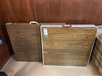 Two vintage folding tables folded and standing upright, showing woodgrain laminate tops and carrying handles.