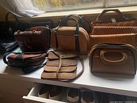 Photo showing assortment of nine purses on a white shelf with a window behind, varying in materials (leather, man-made), colors, and shapes.