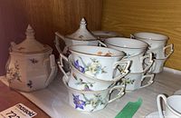 Stacked set of 4 cups and 4 saucers with sugar bowl and teapot in the background, all white with floral patterns and gold rims.