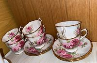 Group of six Royal Albert American Beauty teacups and matching saucers arranged on a shelf showing floral pattern and gold rim detail.