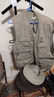 Photo of one Tilley beige vest on hanger with multiple pockets visible, and one Tilley hat on chair below.