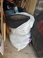 Stack of four tires wrapped partly in a white protective bag, showing their sidewalls and tread tops, stored in a garage with surrounding items.