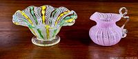 Two pieces displayed side-by-side on wood surface: a ruffled footed dish in clear latticinio with colorful swirls, and a pink miniature pitcher/vase with latticinio threads and clear handle.
