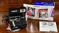 Front angled view showing Polaroid Supercolor camera with original box and manual in background on wood surface.