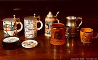 Wide view showing six beer steins including three with pewter lids, one pewter pitcher, and a set of coasters near them on a wooden surface.