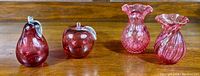 Photo showing two cranberry glass vases with different designs and two cranberry glass fruit figurines (pear and apple) on a wooden surface.