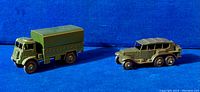 Two vintage military Dinky Toys on blue background: a 6-wheel reconnaissance vehicle and a covered wagon truck.
