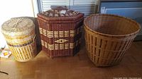 Three wicker baskets placed on a wooden floor against drawers. Two round baskets and one hexagonal corner basket with lids are shown.
