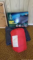 Photo showing boxed Woods queen size double high air bed with a red sleeping bag compression sack and black fabric item on carpet, in front of wood panel wall.