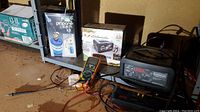 Photo showing three battery chargers and an Extech digital multimeter with test leads connected, all on a metal shelf with concrete background.