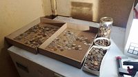 Two cardboard trays, plastic container and jar filled with loose coins on a work surface.