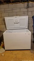 White Kenmore chest freezer with lid open, showing interior and wire basket, placed in basement storage area.