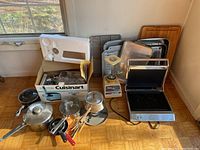 Photo showing various pots with lids, a boxed Cuisinart meat grinder, a blender, a sandwich press, baking trays, and cooking utensils on floor by window
