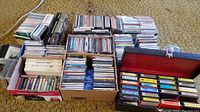 Overview of boxed collection of 8 track tapes, cassettes, and CDs arranged on carpeted floor.