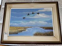 Full view of the framed painting showing Canada geese flying over marsh with water and reeds, snowy mountains in background. Frame shows staining and wear.