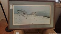 Full view of the framed art print depicting wolves in the snow, showing the entire piece on a stand by a window.