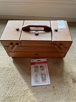 Closed wooden sewing box with handle, along with unopened pack of safety pins placed below it.